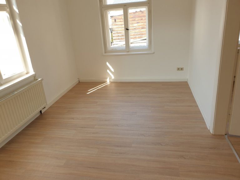 brauner PVC-Belag, weiße Wände, 2 große Fenster, weißer Heizkörper