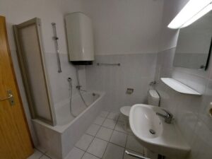 weiße Fliesen an Wand und Boden, Badewanne mit Duschwand, Waschbecken, Spiegel, WC, Warmwasserspeicher