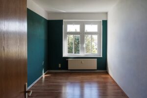 brauner Parkettboden, zwei weiße und zwei dunkelgrüne Wände, weißes Fenster, weißer Heizkörper unter Fenster