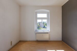zwei weiß und eine grau gestrichene Wand, brauner Parkettfußboden, weißes Fenster, unter Fenster weißer Heizkörper