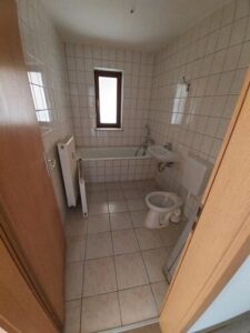 weiße Wand- und Bodenfliesen, braune Holztür, Fenster mit braunem Holzrahmen, davor Badewanne weiß, weißes WC stehend, Waschbecken weiß, weißer Heizkörper