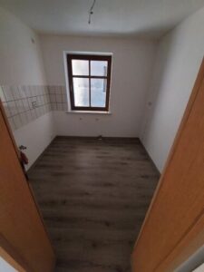 grauer Laminatboden, braune Holztür, braunes Holzfenster, weiße Wände, weißer Fliesenspiegel links