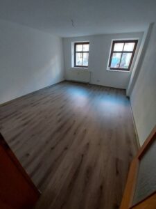 grauer Laminatboden, weiße Wände, braune Holztür, 2 braune Holzfenster, weißer Heizkörper unter Fenster