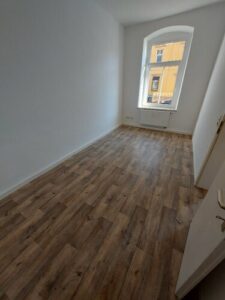 Zimmer mit braunem Parkettboden und einem weißen Fenster. Wände sind weiß gestrichen.