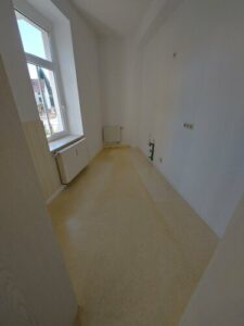 Küche mit beigefarbenem Laminatboden und weißen Fenster. Wände weiß gestrichen. weißer Heizkörper unter Fenster. Anschlüsse auf der rechten Wand vorhanden. Fenster gegenüber.