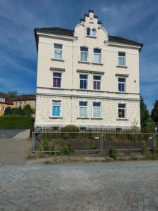 Ansicht des Gebäudes von außen (straßenseitig). Links vom Objekt ist der Zugang zum Haus, Parkplatz und Garten. Fassade ist hellgestrichen.