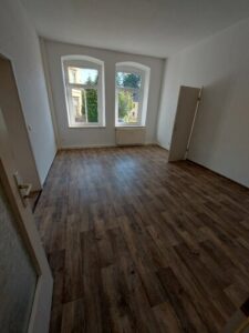 Zimmer mit braunem Parkettboden und zwei weißen Fenstern. Wände weiß gestrichen.