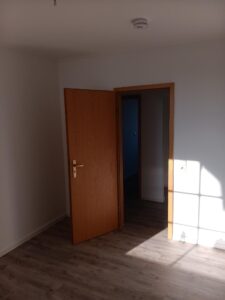 brauner Parkettfußboden, weiße Wände, braune Holztür, Rauchwarnmelder an Decke