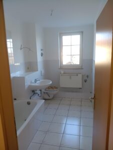 weiße Wand- und Fußbodenfliesen, braune Eingangstür, weiße Badewanne, weißes Waschbecken, Fenster, weißer Heizkörper unter Fenster, WC