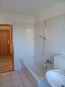 weiße Wand- und Bodenfliesen, weiße Badewanne, Dusche, weißes Waschbecken, braune Holztür