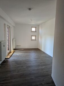 Zu sehen ein rechteckiger leerer Raum mit weißen Wänden, holzfarbenen Boden und einer Balkontür und zwei kleinen quadratischen Fenstern