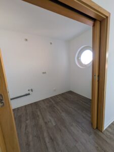 Zu sehen ein leerer Raum mit weißen Wänden, rundem Fenster und holzfarbenen Boden
