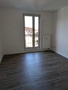 Zu sehen ist ein Zimmer mit weißen Wänden, einer Balkontür und holzfarbenen Boden