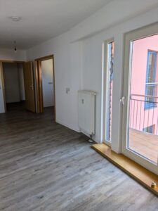 Zu sehen ein leerer Raum mit weißen Wänden, holzfarbenen Boden und 2 holzfarbenen Zimmertüren sowie einer Balkontür