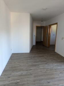 Zu sehen ein rechteckiger leerer Raum mit weißen Wänden, holzfarbenen Boden und 2 holzfarbenen Zimmertüren