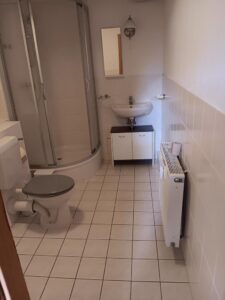 Bad mit Dusche im hinteren Eck, Waschbecken, WC, Waschmaschine, weiße Fußboden- und Wandfliesen