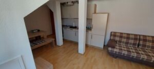 brauner Laminatboden, weiße Wände, Einbauküche mit Kühlschrank, Herd, Spüle, Sitzecke, Couch, Fenster, zwei Säulen