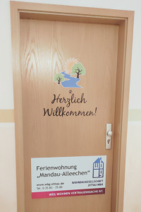 Wohnungseingangstür , wo ein das Logo der WBG Zittau zu sehen ist und mit folgendem Schriftzug versehen ist: Herzlich Willkommen - Ferienwohnung "Mandaualleechen", Telefon: 03583-7500