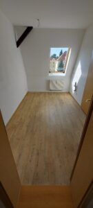brauner Laminatboden mit Maserung, weiße Wände, Holzdachflächenfenster, weißer Heizkörper, Anschlüsse für Küchengeräte