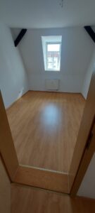 hellbrauner Laminatboden, weiße Wände, dunkle Holzbalken an den Dachschrägen, weißes Fenster, unter dem Fenster ein weißer Heizkörper