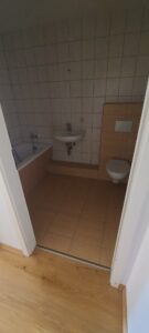 weiße Wandfliesen, braune Fußbodenfliesen, WC hängend, Aufputzspülkasten, Waschbecken, Badewanne mit Armaturen