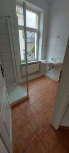 weißer Wandfliesenspiegel, rötlichbraune Fußbodenfliesen, Dusche, Waschbecken, Fenster weiß, Heizkörper weiß unter Fenster