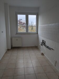 Rechteckiger Raum mit beigefarbenen Bodenfliesen, einen hellen Fliesenspiegel an der rechten Wand und vorne ein Fenster mit einem Heizkörper