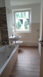 Bad mit holzfarbenen Bodenfliesen, links befindet sich eine Wanne, ein Waschbecken, 1 WC und rechts ein Heizkörper. Die Wände sind hell.