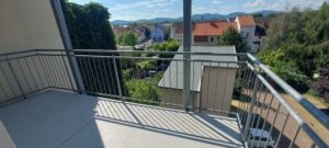 Balkon, grauer Plattenboden, graues Stahlgeländer, Ausblick ins Gebirge (Berge, grüne Bäume, Nachbarhaus)