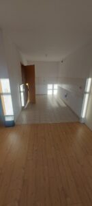 brauner Laminatboden, Durchgang zur Küche, weißer Fliesenboden angrenzend, weiße Wände, Fliesenspiegel in Küche, braune Holztür Richtung Flur, weißer Heizkörper neben Tür in Küche