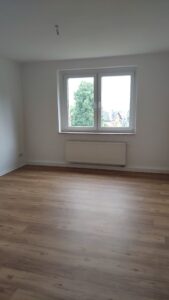 rechteckiger Raum mit holzfarbenen Fußboden und weißen Wänden. Vorne ein Fender und ein weißer Heizkörper