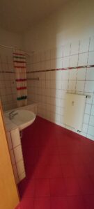 weiße Wandfliesen mit roter Bordüre, roter Fliesenboden, weißes Waschbecken links, weiße Badewanne, weißer Heizkörper, gemusterter Duschvorhang